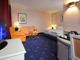 Einblick in das Zimmer mit orangefarbener Eckcouch vor dem Doppelbett auf blau gemustertem Teppichboden 